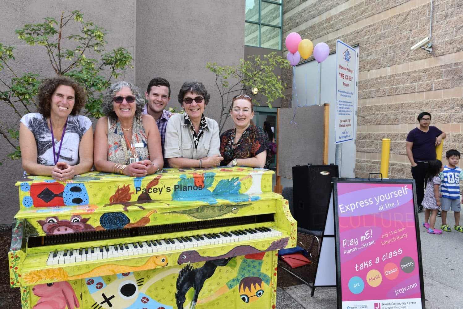 Five smiling volunteers lean shoulder-to-shoulder over a chartreuse folk-art piano at a community-centre launch party.