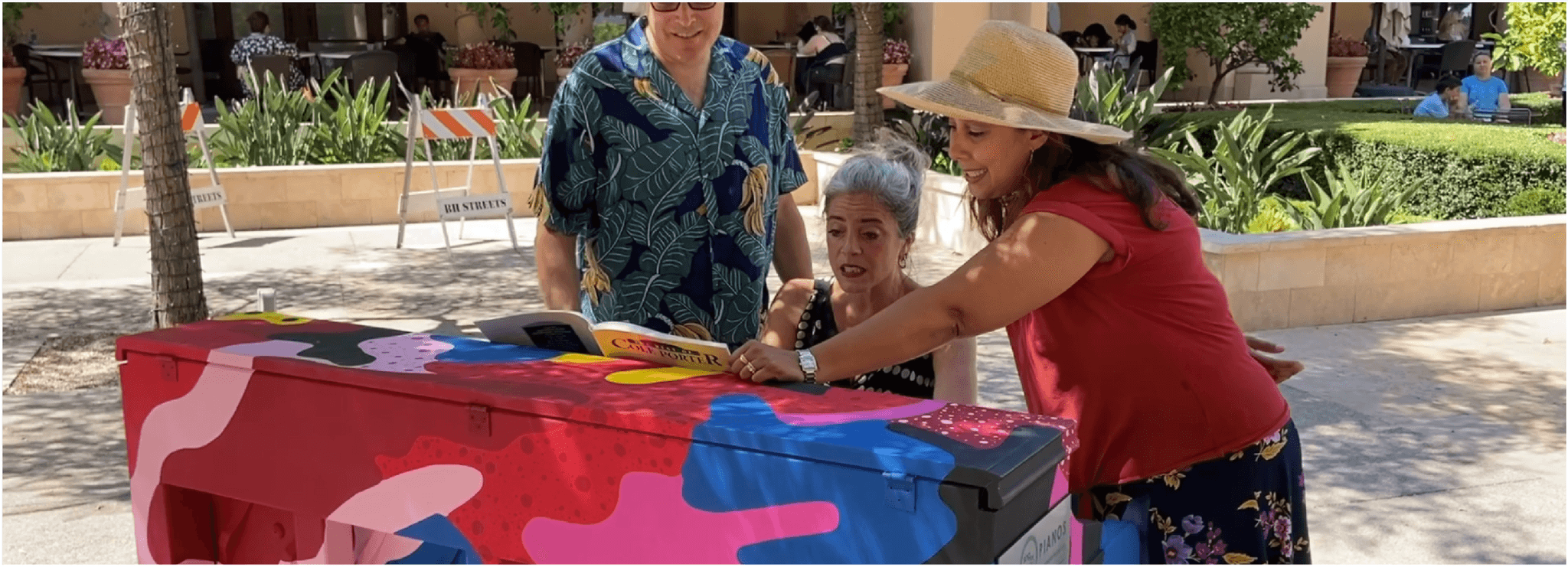 Three friends huddle over a Cole Porter songbook on a pink-and-blue camo piano in a palm-lined courtyard.