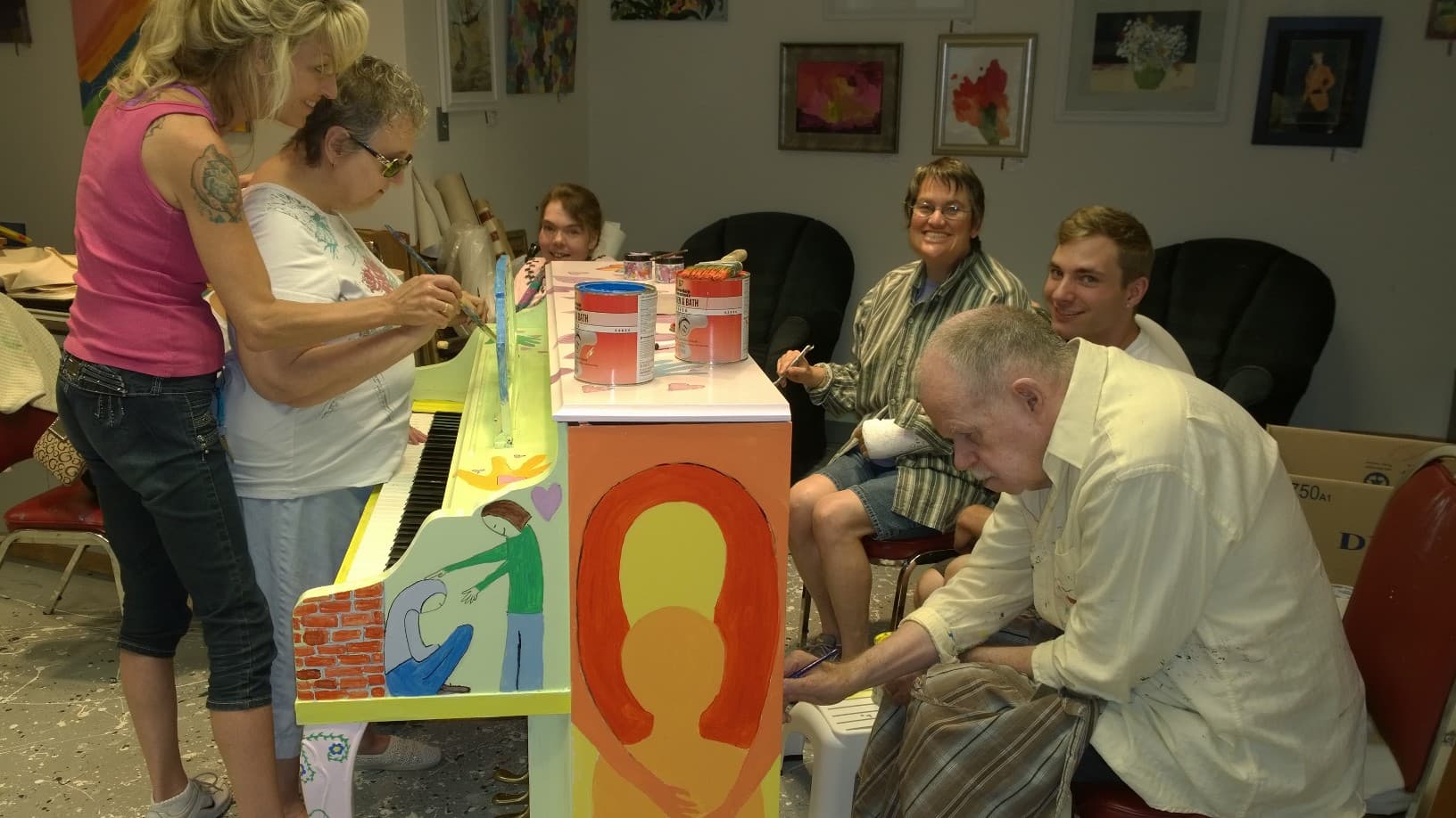Artists crowd a studio to hand-paint a piano, paint cans balanced on the lid.
