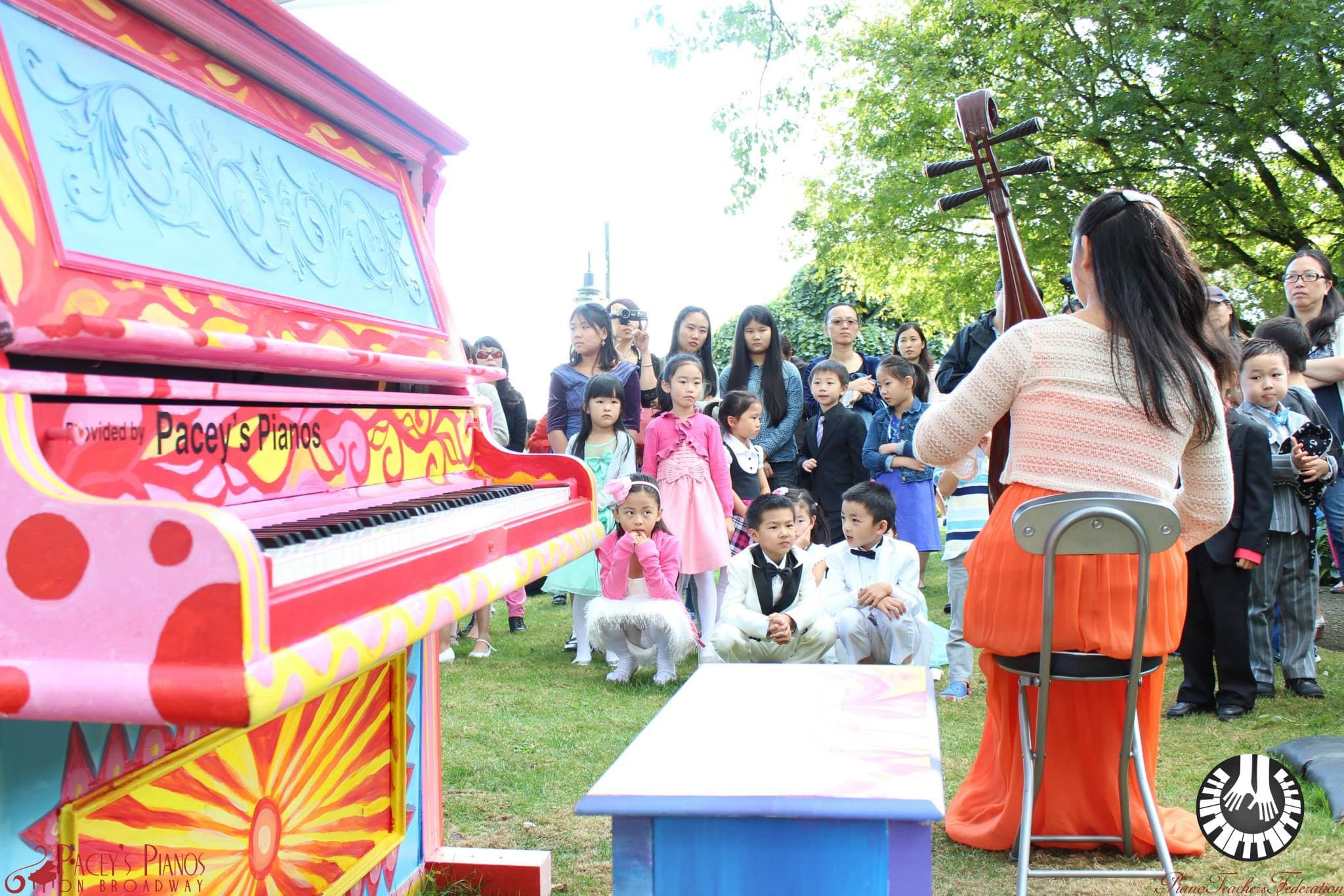 A hot-pink psychedelic piano anchors a park recital as a pipa player in orange performs for rows of children.