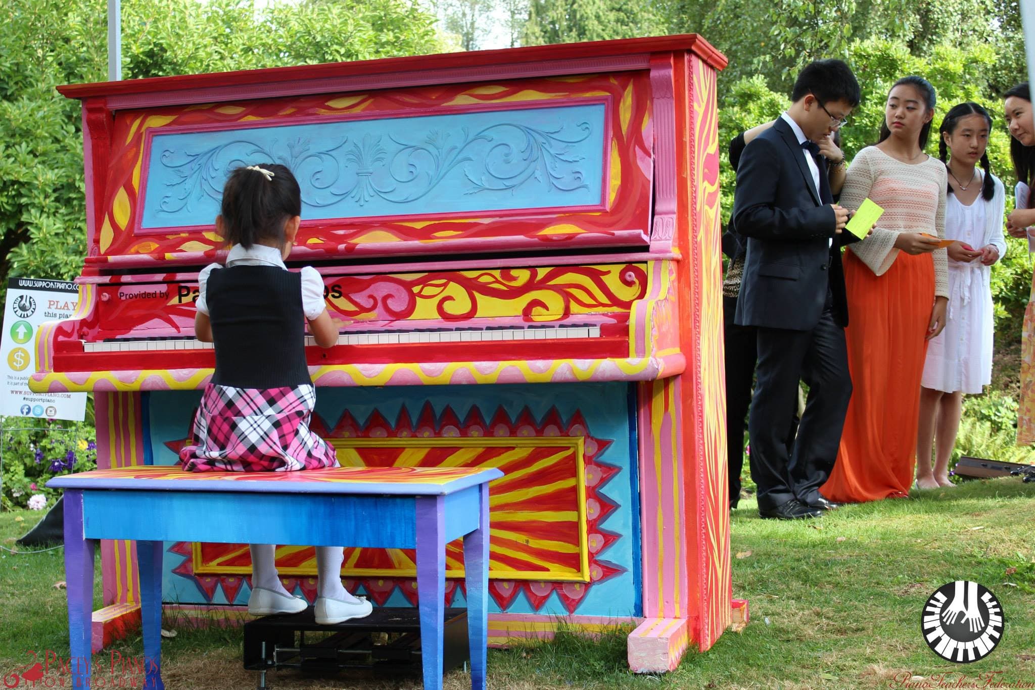 A small girl stands on a stool to reach the keys of a towering red baroque-painted upright in a garden.