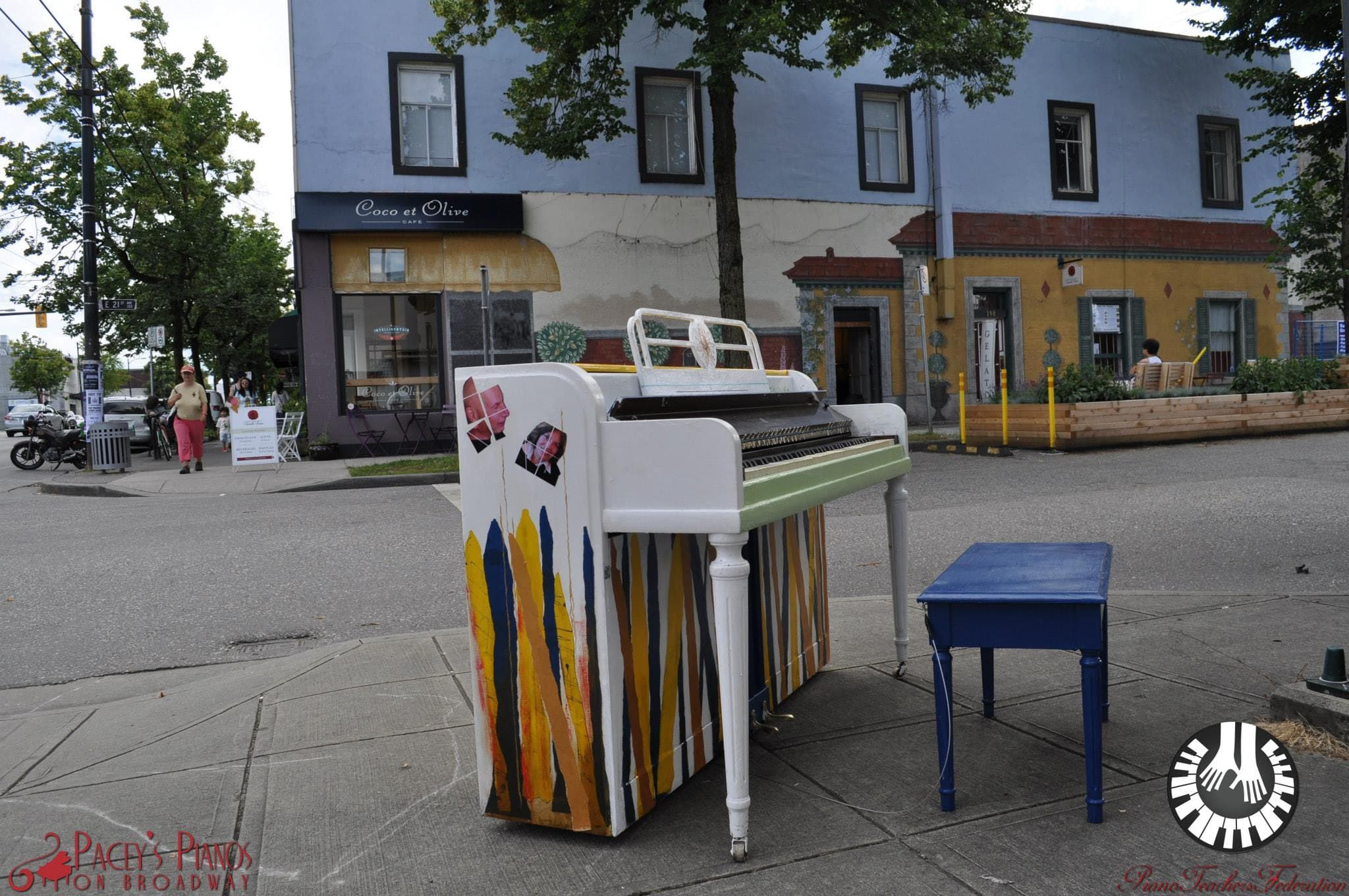 A white piano striped with ribbons of yellow, blue and orange waits with a cobalt bench on a quiet cafe corner.