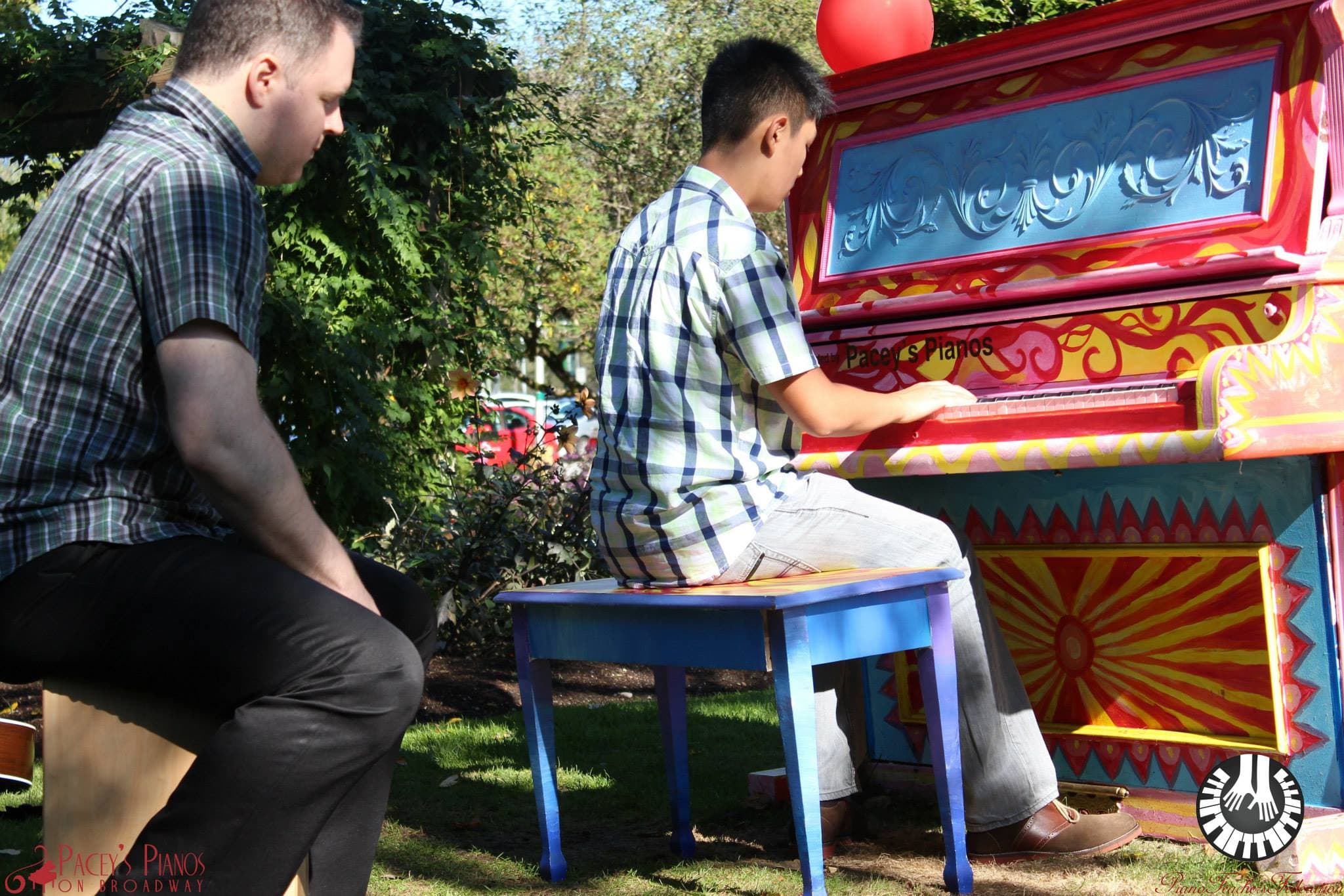 A teenager plays the red baroque sunburst piano in a garden while a man on a cajón keeps time.