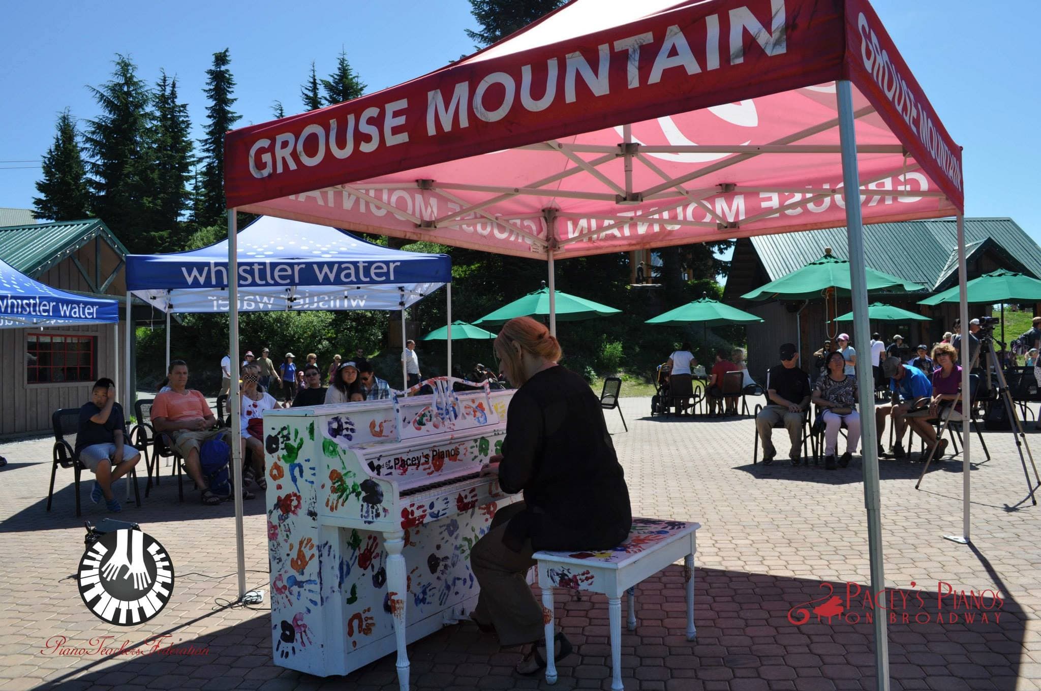 A pianist plays a rainbow-handprint piano beneath a red canopy on the Grouse Mountain plaza, crowd seated all around.