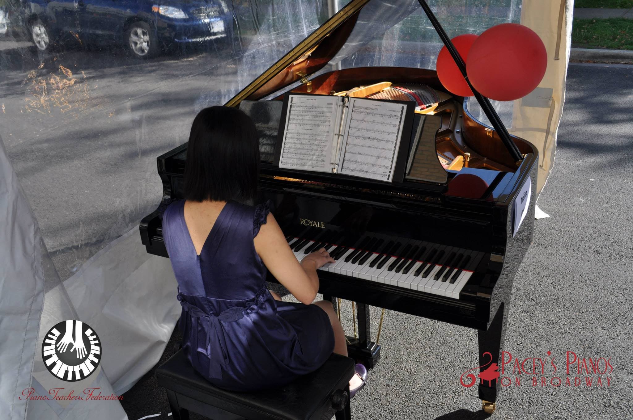 A woman in a violet satin dress plays a gleaming black baby grand under a clear-walled tent, red balloons over the open lid.