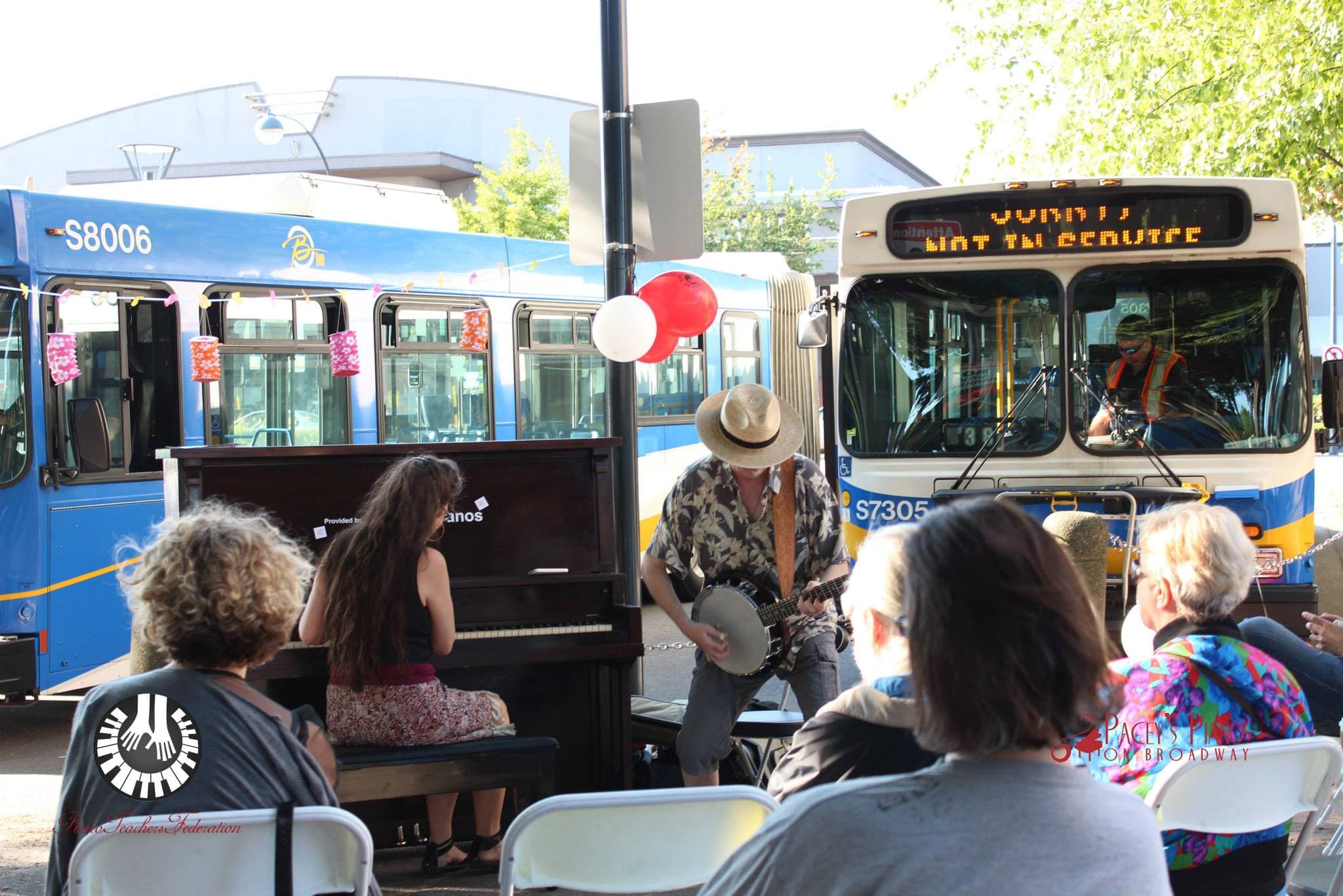 Piano and banjo jam at a lantern-strung bus exchange while an audience in folding chairs listens.