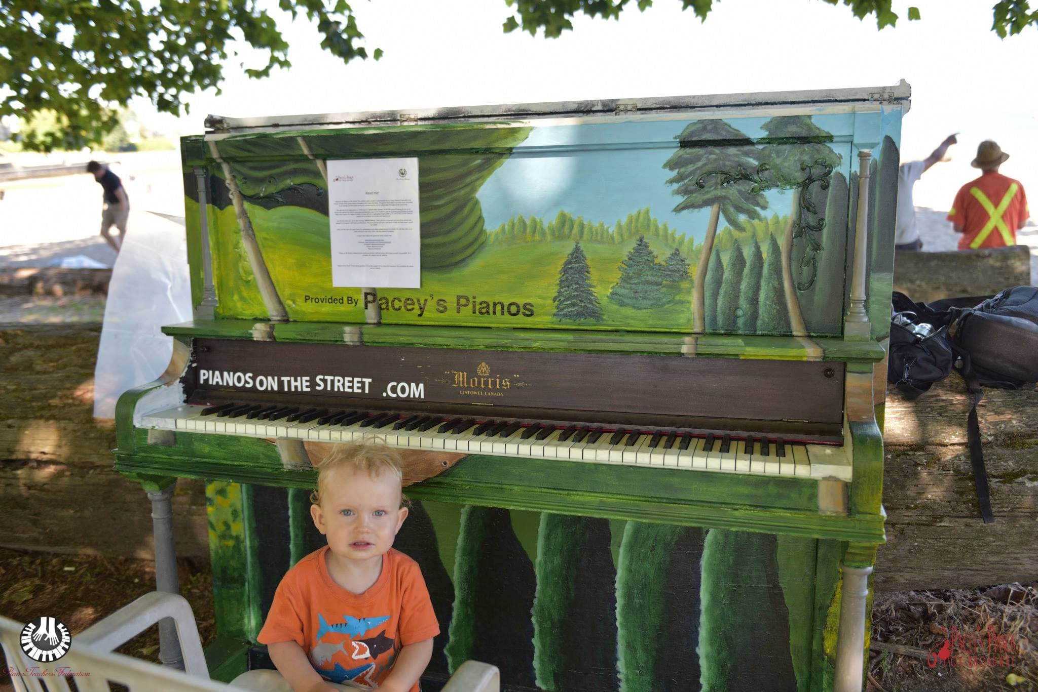 A toddler in an orange shark t-shirt reaches the keys of the forest-mural upright, parked against beach driftwood.