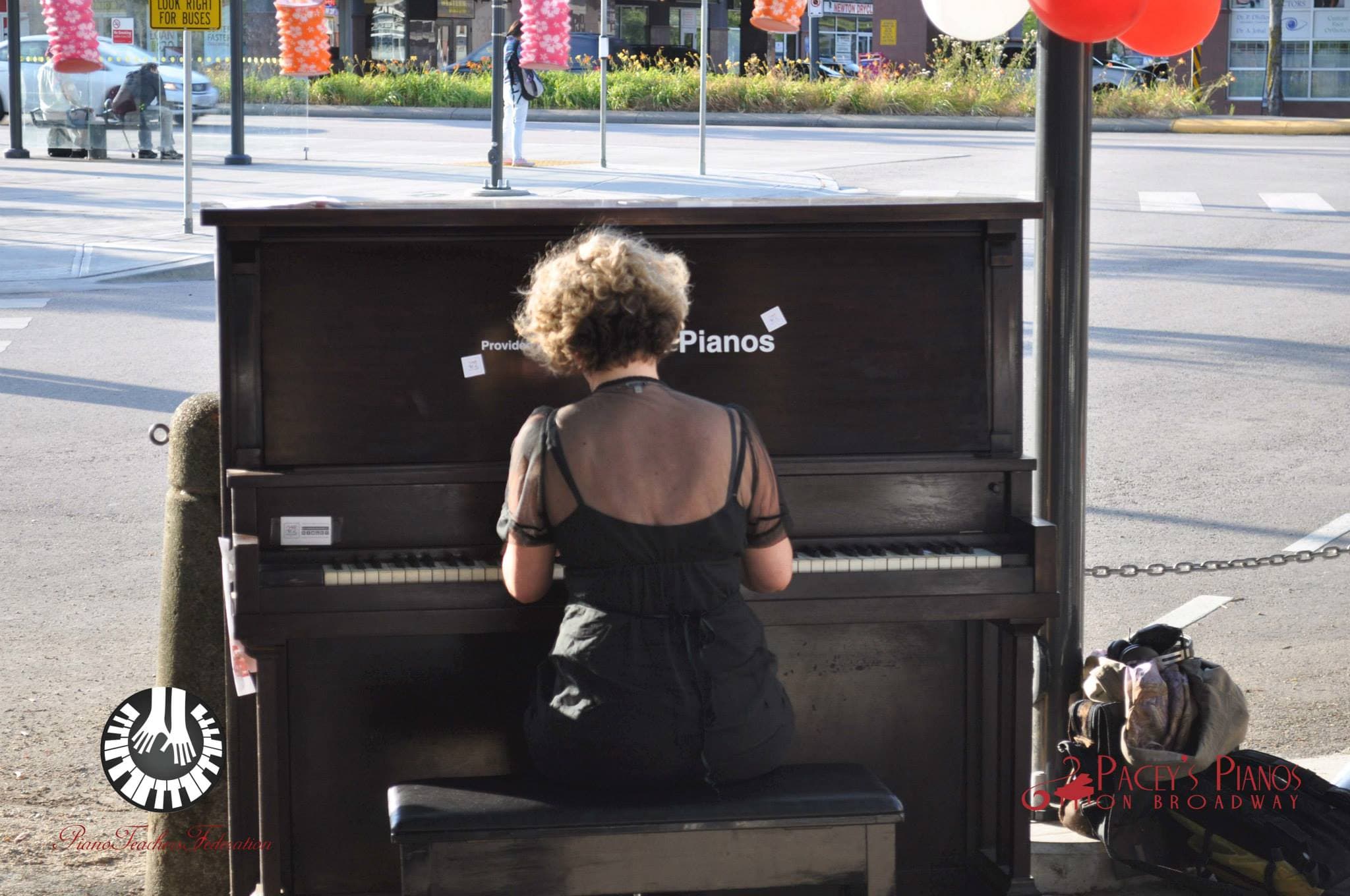 In golden late-afternoon light, a woman plays a dark-wood upright at a crosswalk corner, red balloons overhead.