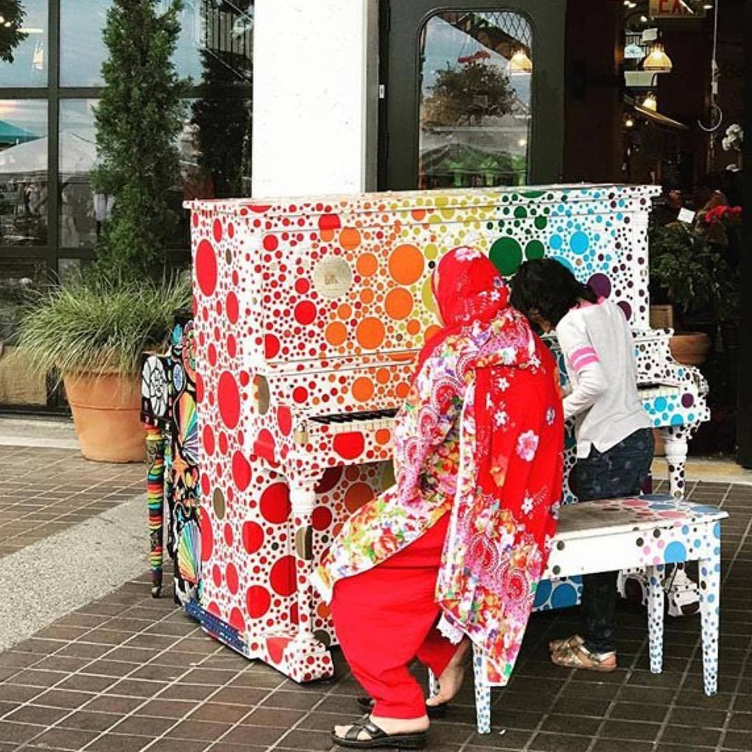 A mother in red florals and a girl in white play a rainbow polka-dot piano together outside a mall entrance.