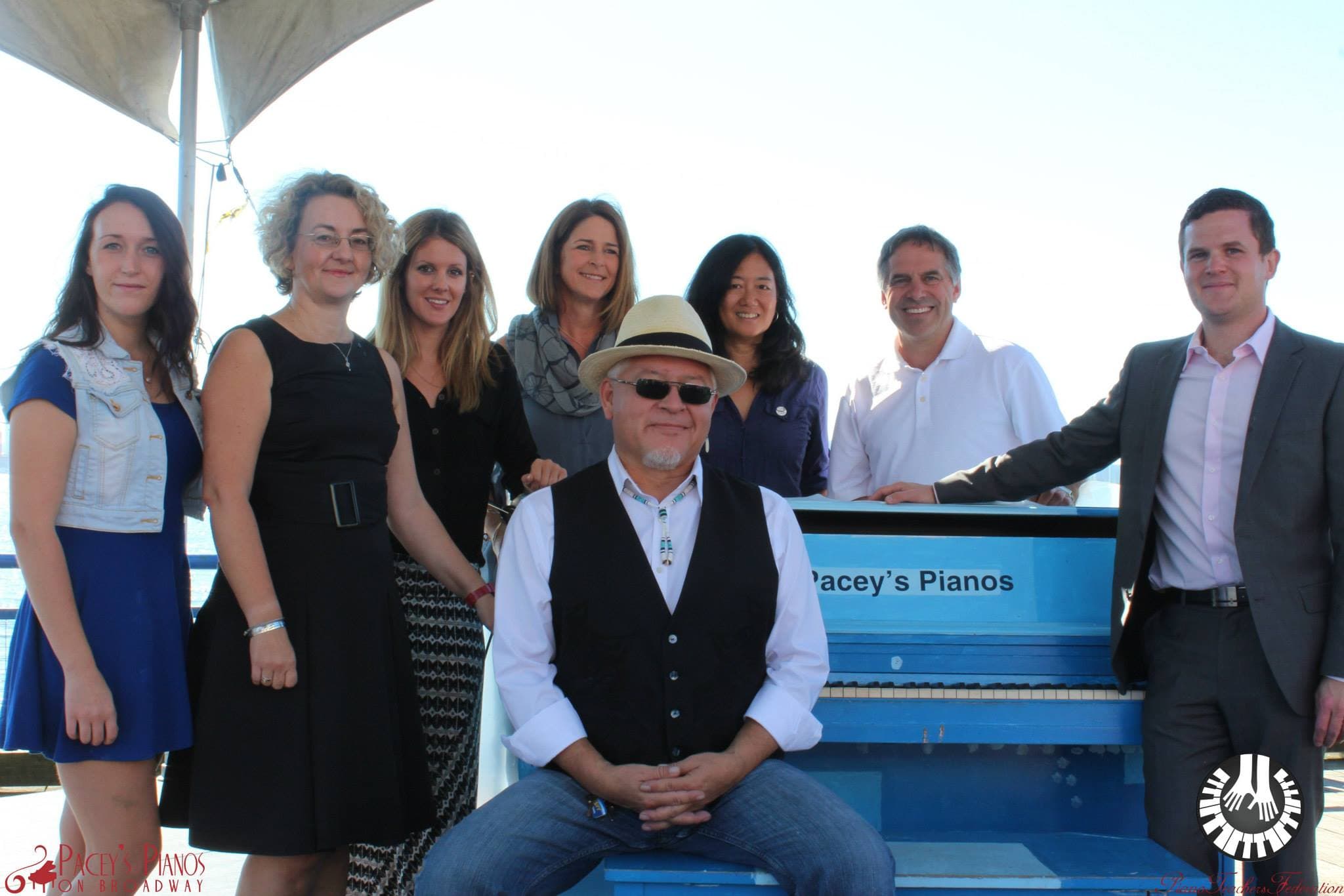 Organizers and supporters pose around a powder-blue Pacey's Pianos upright under a white sail canopy on a waterfront pier.