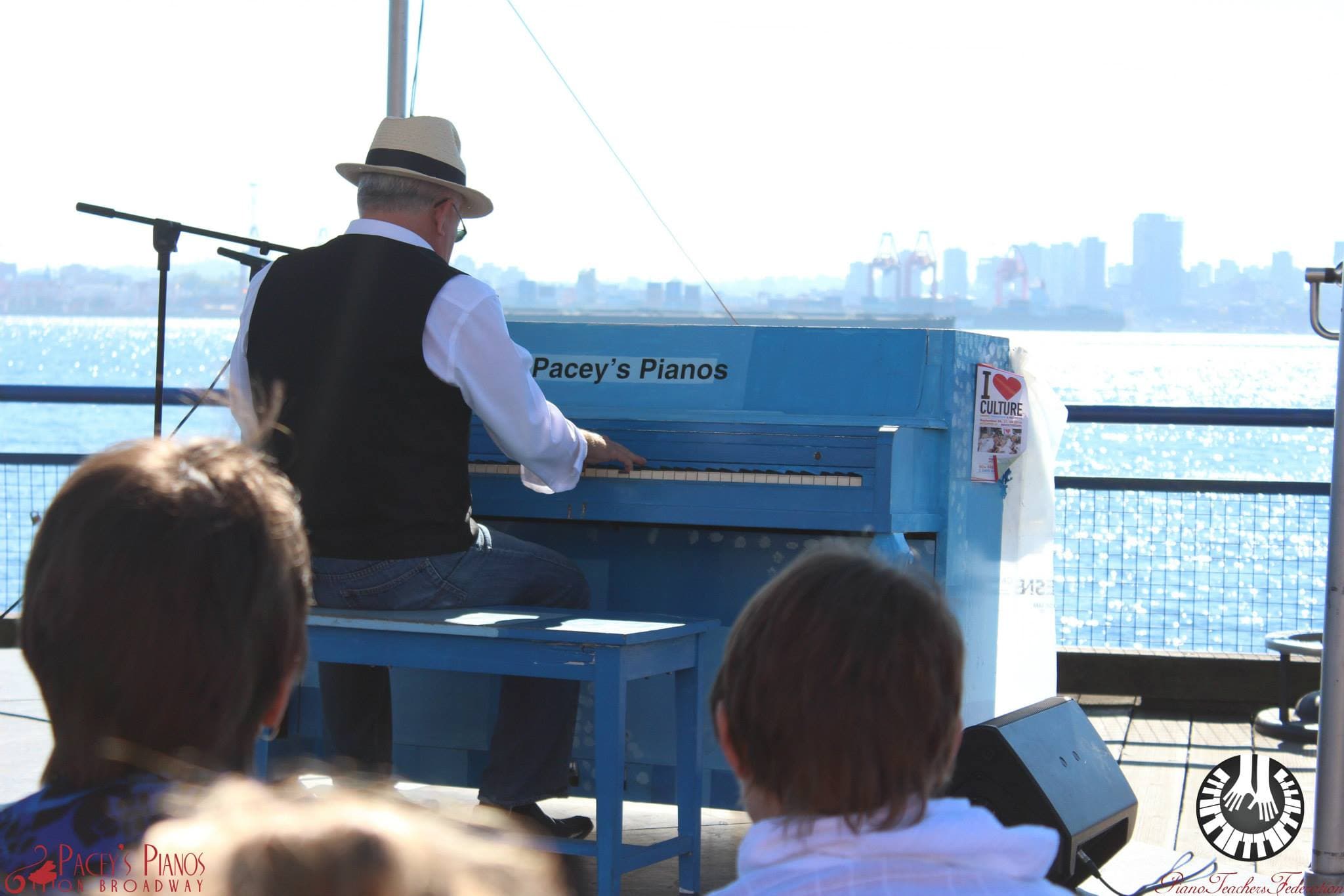 A man in a straw fedora plays a sky-blue piano at the edge of a pier, the Vancouver skyline hazy across the water.