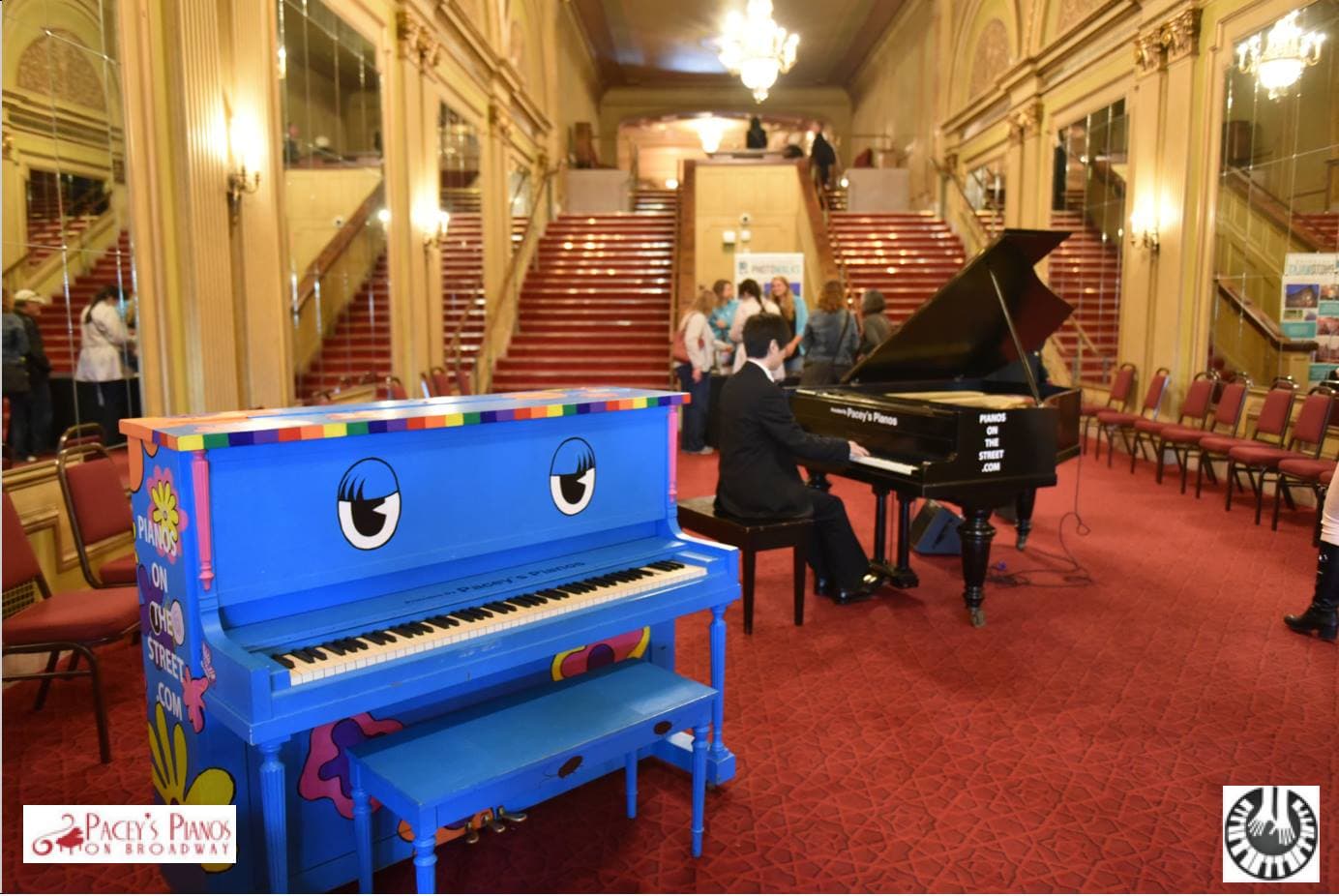 A cartoon-eyed blue upright grins in a chandelier-lit theatre lobby with red-carpeted grand staircases.