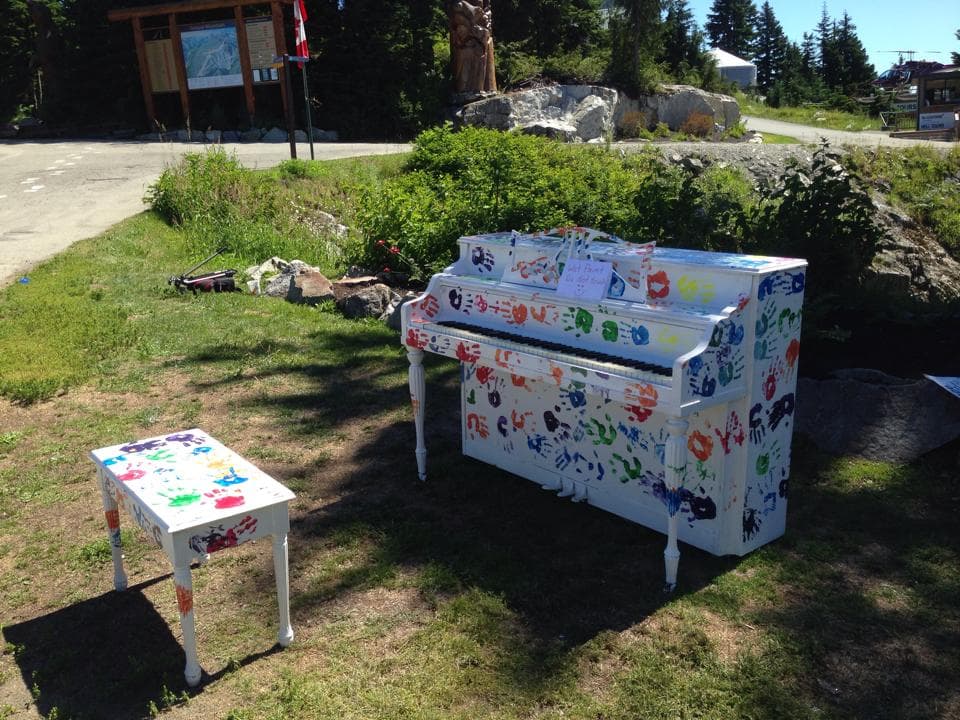 A white piano stamped with rainbow handprints on trailhead grass at Grouse Mountain, wet-paint sign still taped up.