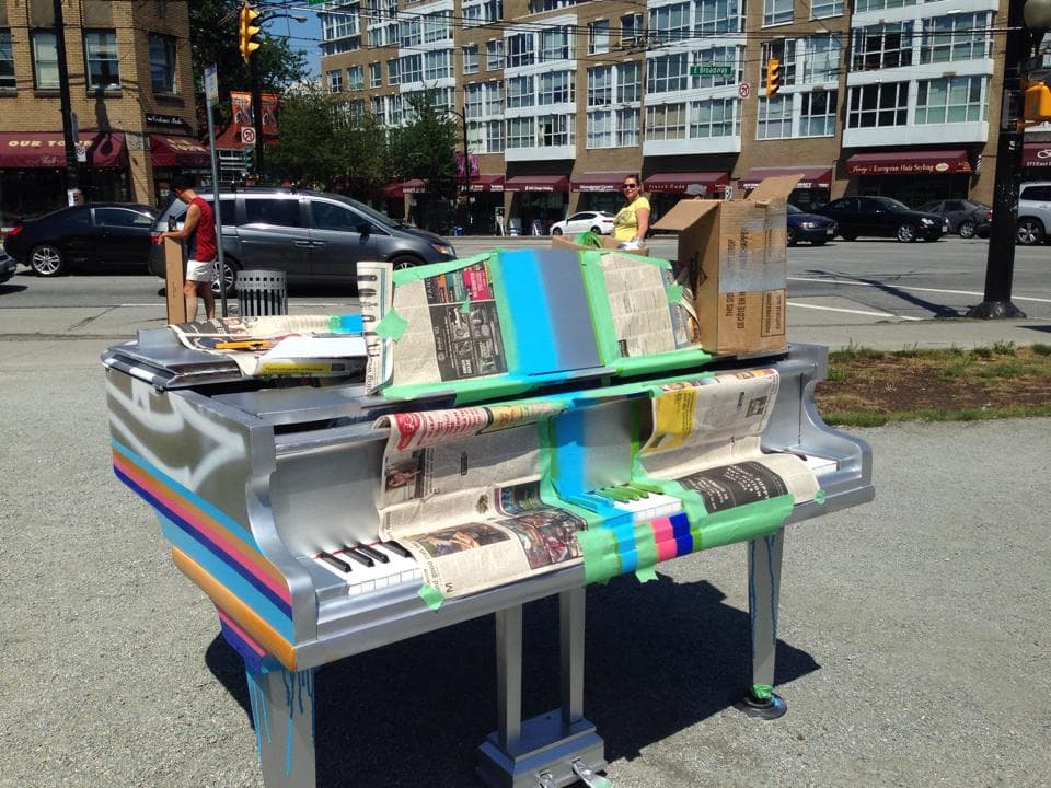 A silver baby grand mid-makeover, panels masked with newspaper and tape while fresh candy stripes dry.