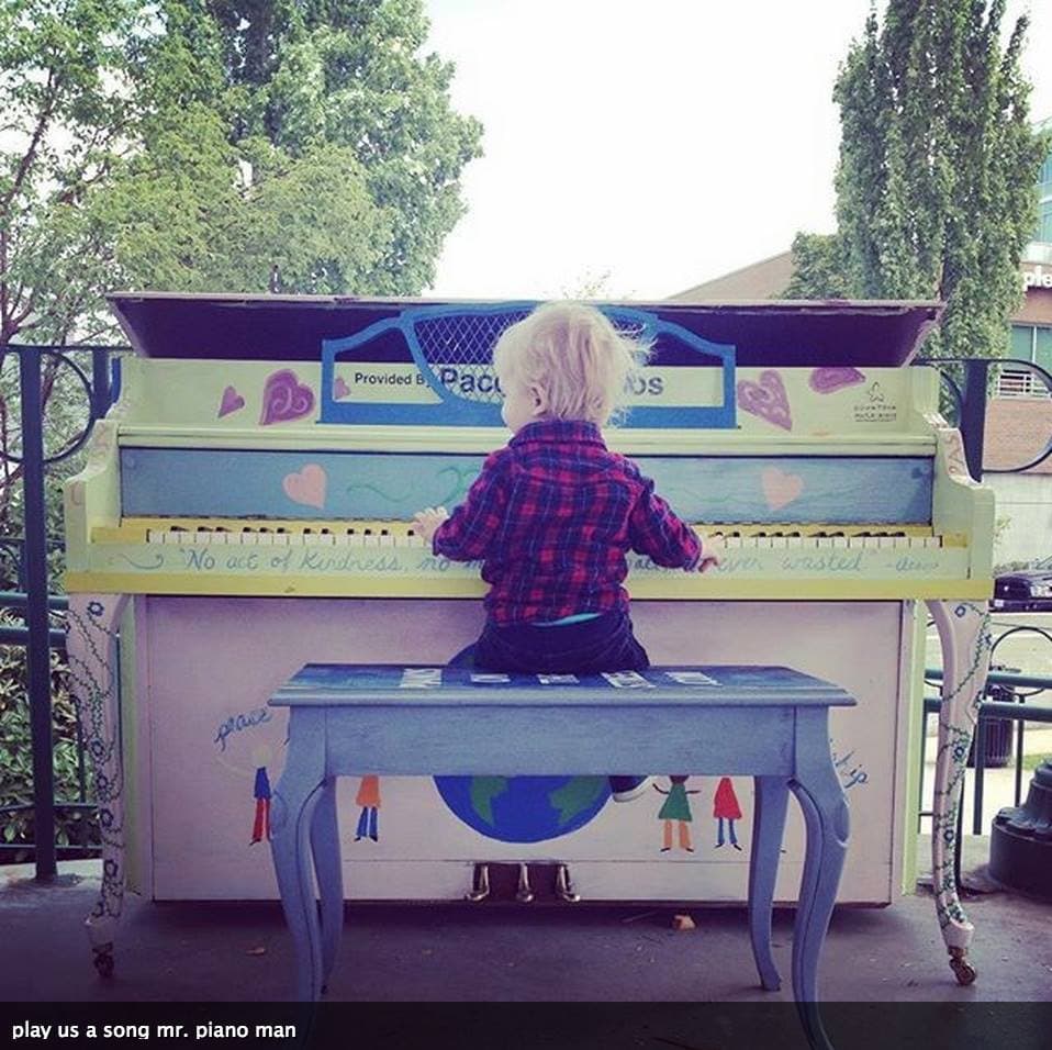 A toddler kneels on a painted bench, arms stretched across the keys of a piano lettered 'No act of kindness is ever wasted'.
