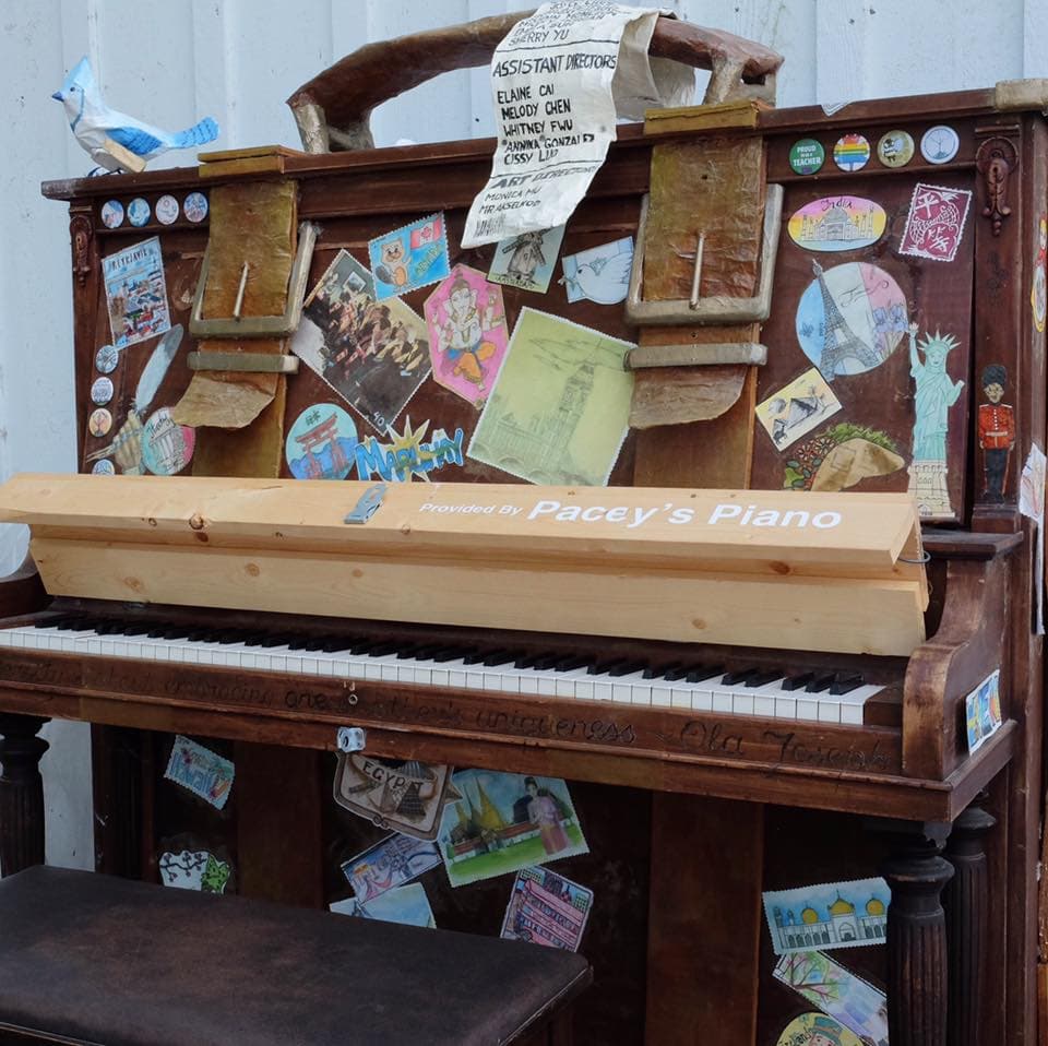 A piano transformed into a giant vintage travel suitcase with leather straps, postcard stamps, and a painted blue jay.