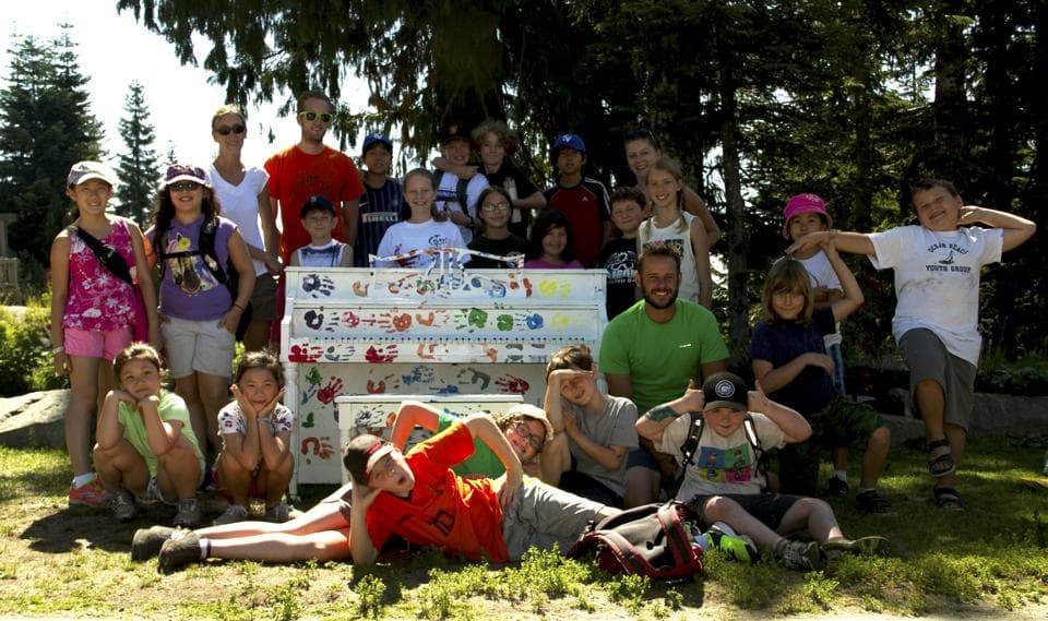 A whole summer camp mugs for the camera around the rainbow-handprint piano in a sun-dappled forest clearing.