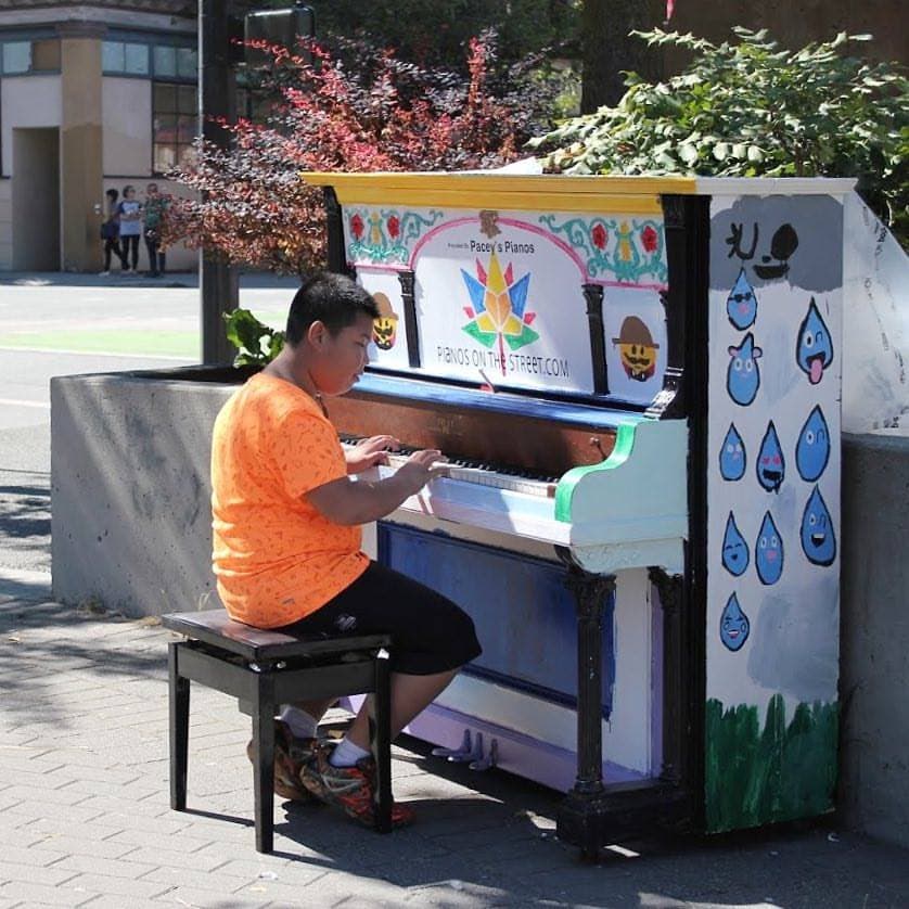 A boy in bright orange plays a folk-art piano with a Canadian maple motif, right on the sidewalk.
