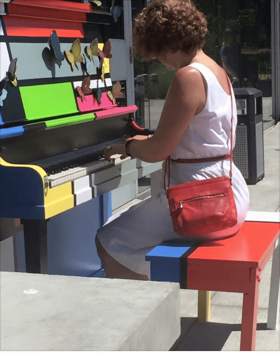 A woman in a white summer dress plays the butterfly-covered Mondrian piano from its colour-block bench.