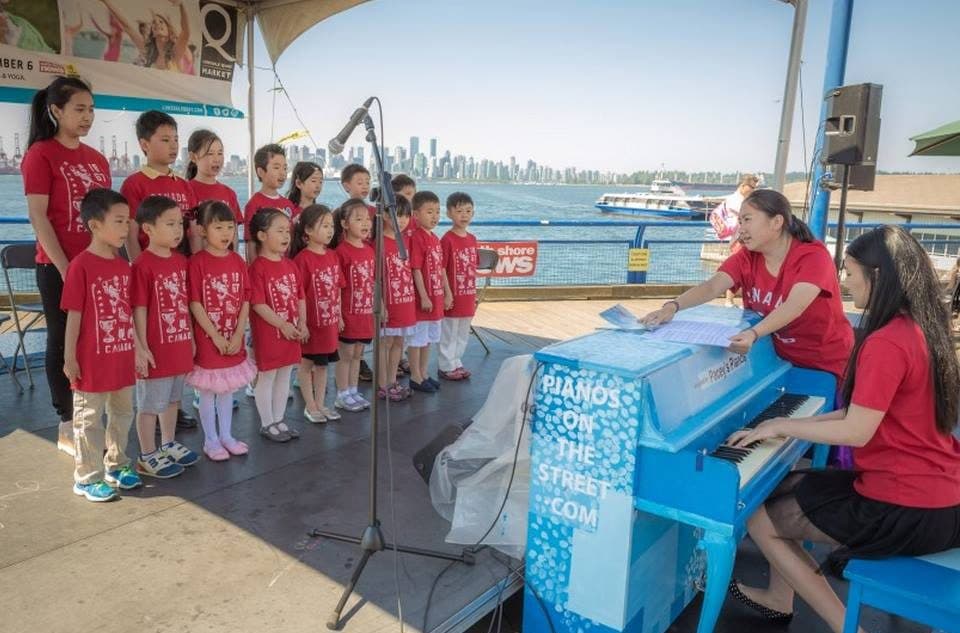 A children's choir in red sings at a North Vancouver pier stage, led from a spotted sky-blue piano, SeaBus beyond.