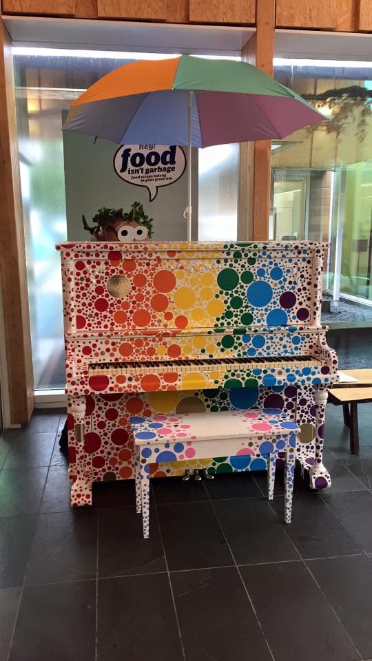 A rainbow polka-dot piano shelters under a rainbow umbrella in a tiled atrium.