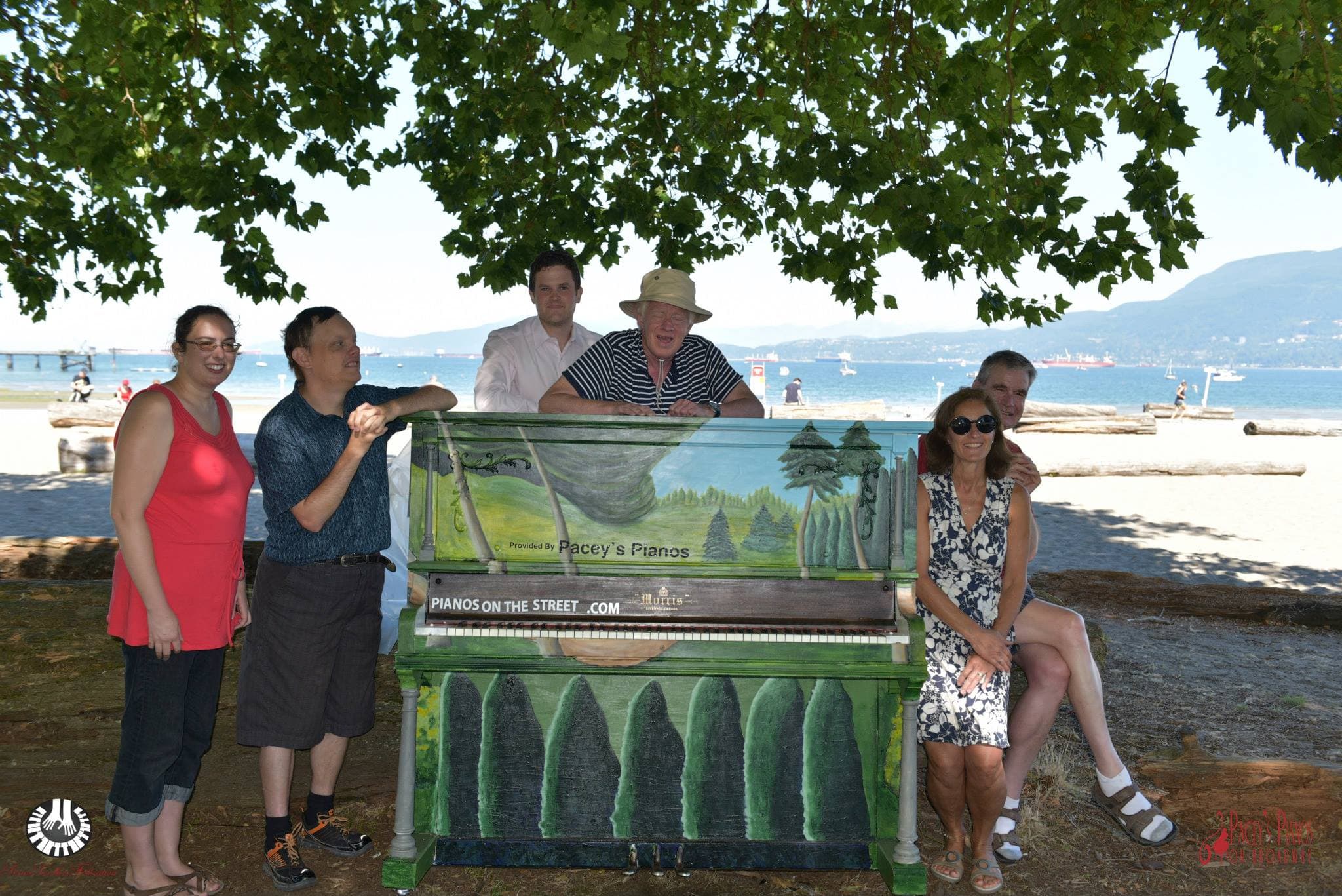 Friends gather around a forest-mural piano under a shady tree at Spanish Banks, mountains across the sparkling water.