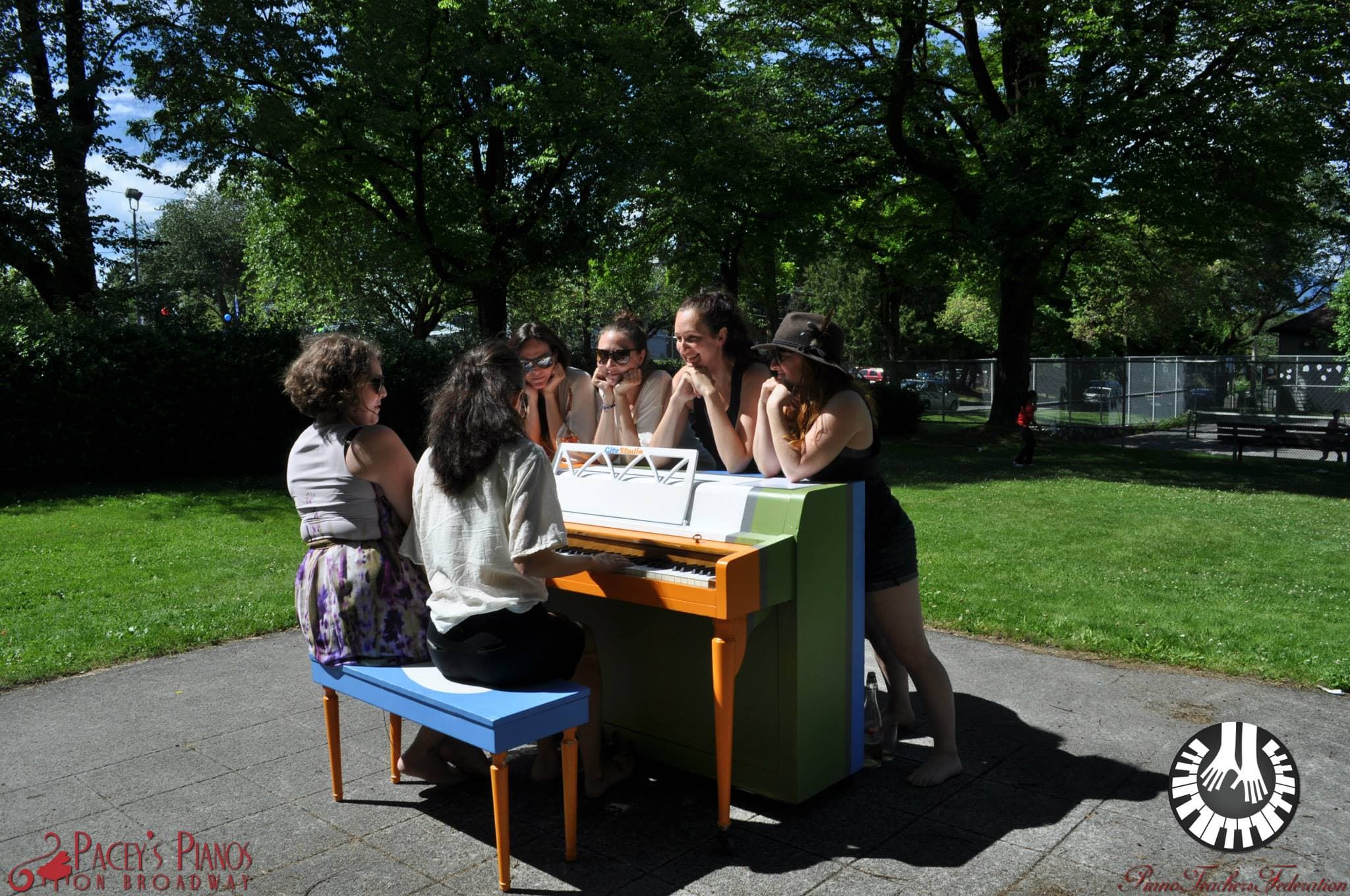 Six young women lean chin-in-hands around an orange-and-green piano in a sunny park circle, listening.