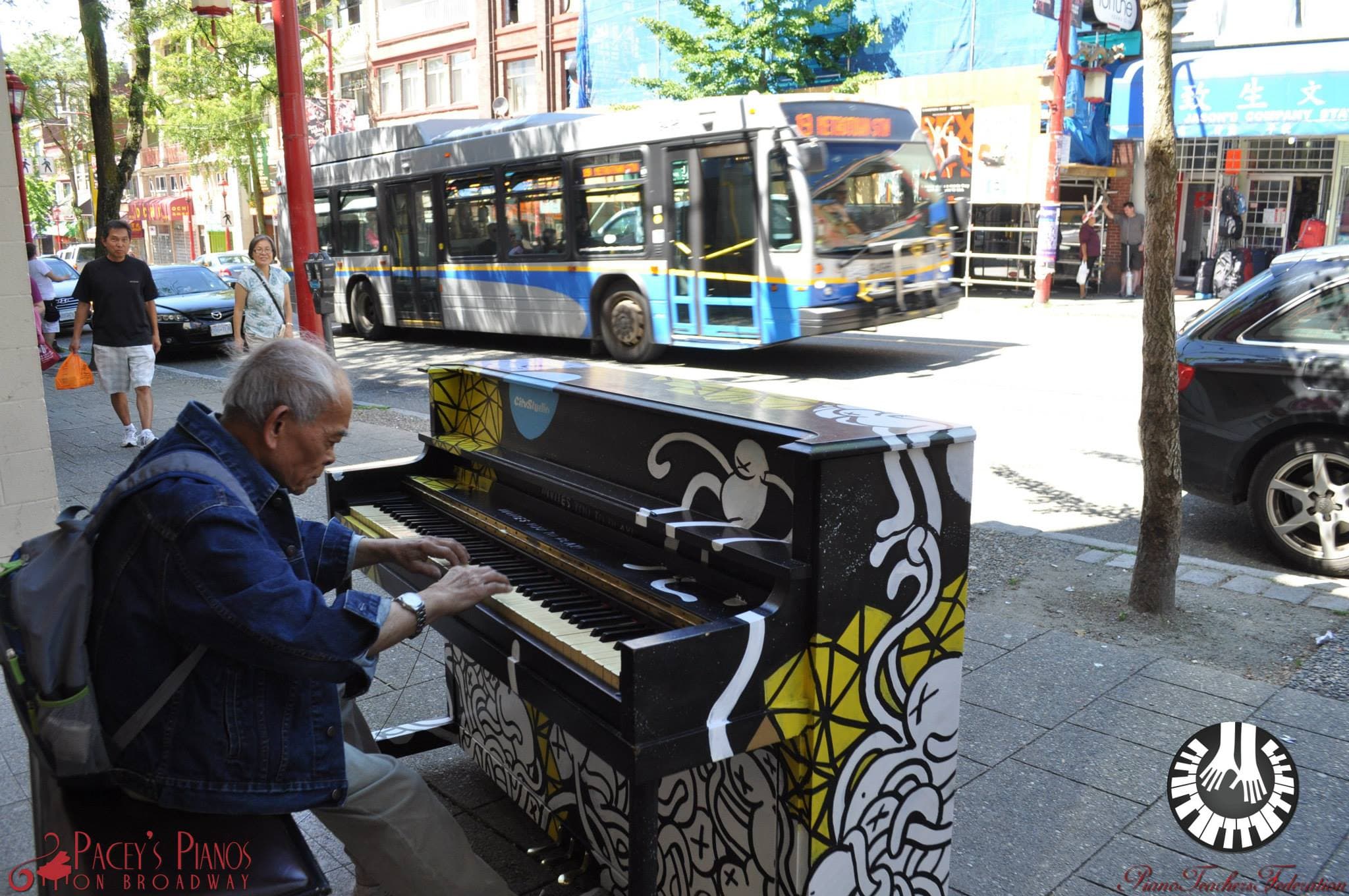 An elderly man in a denim jacket plays a doodle-covered upright on a Chinatown sidewalk as a trolley bus rolls past.